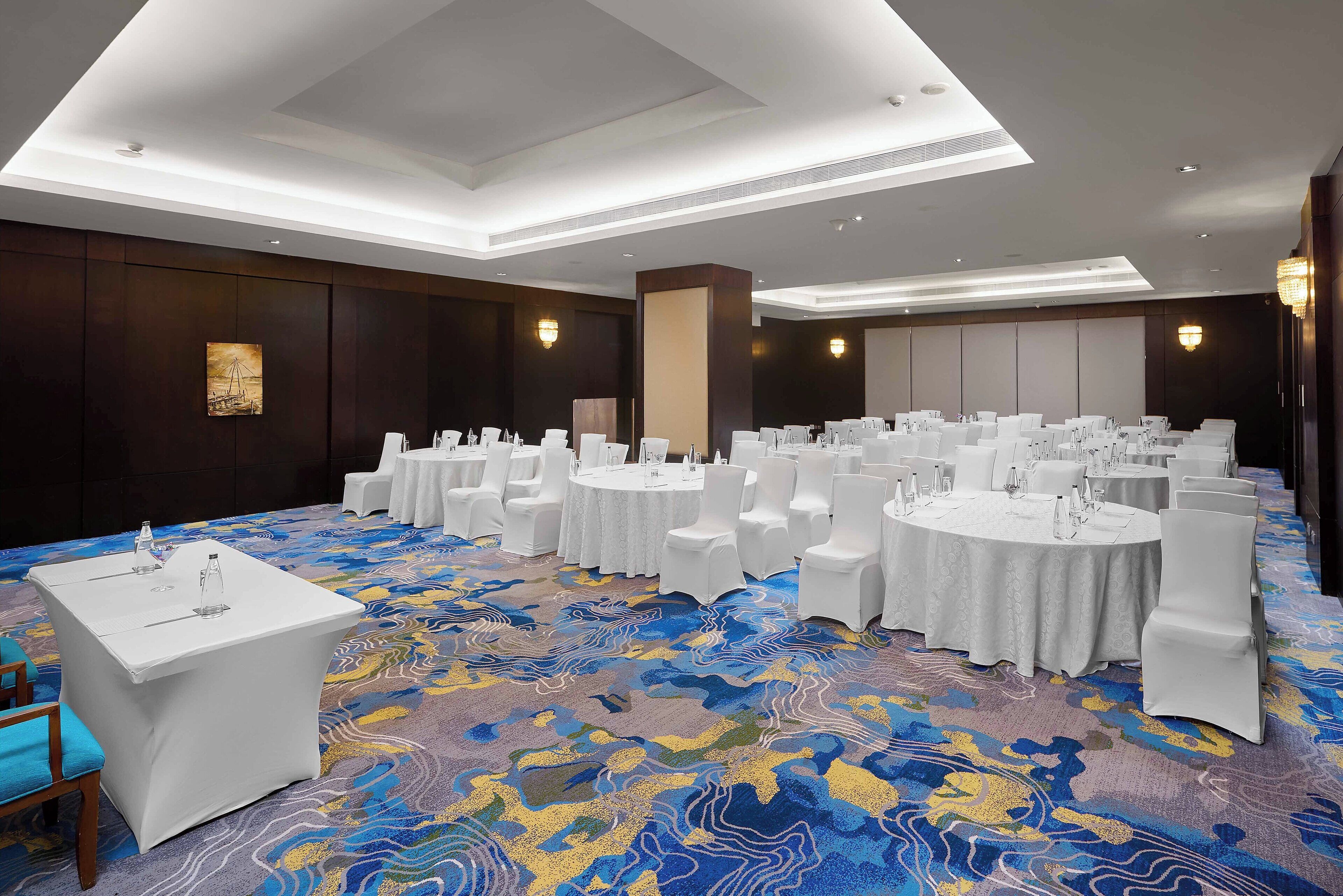 A banquet hall with round tables and white chairs, a patterned carpet, and recessed lighting.