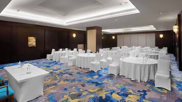 Un salón de banquetes con mesas redondas y sillas blancas, una alfombra estampada e iluminación empotrada.