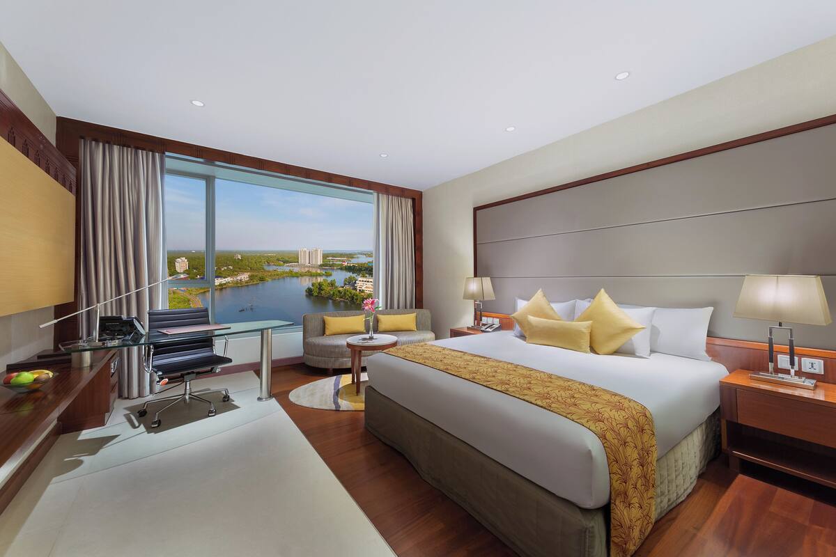 A modern hotel room with a large bed, a desk with a laptop, a sofa, and a view of a cityscape.