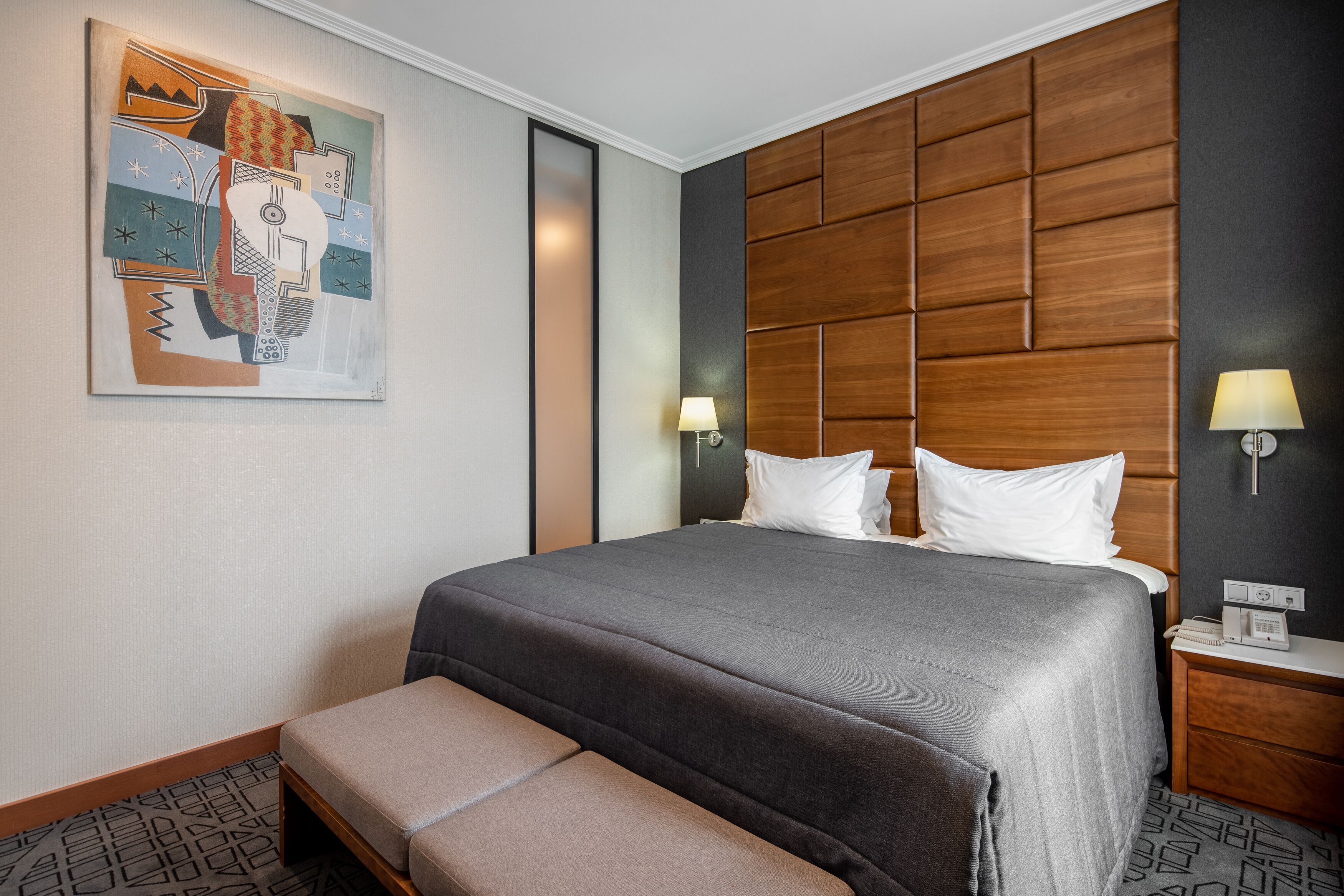 Una habitación de hotel con una cama grande, un banco, cabecero de madera, una lámpara de pared y un cuadro.
