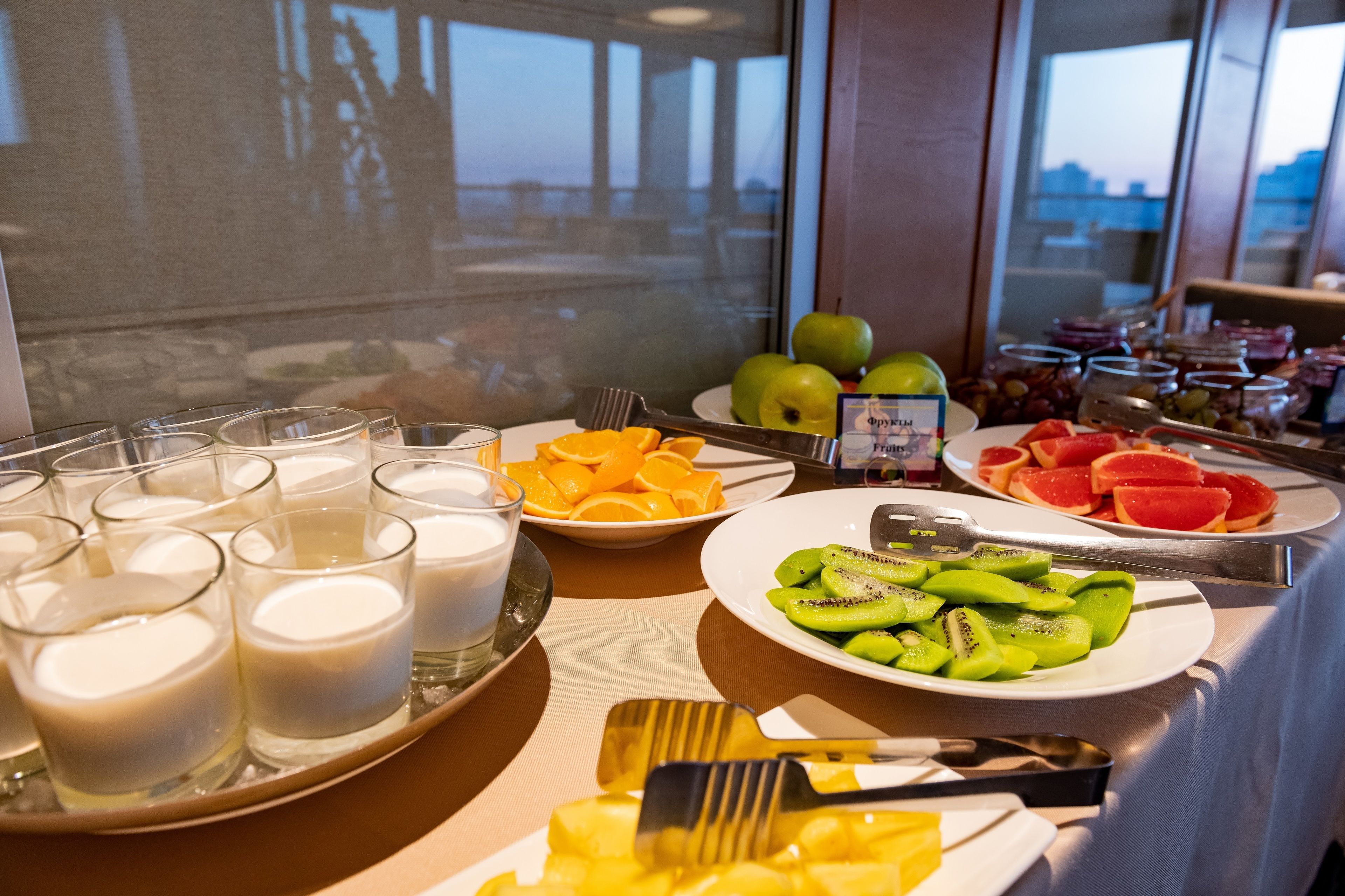 A breakfast buffet with various fruits, yogurt, and a view of the city through large windows.