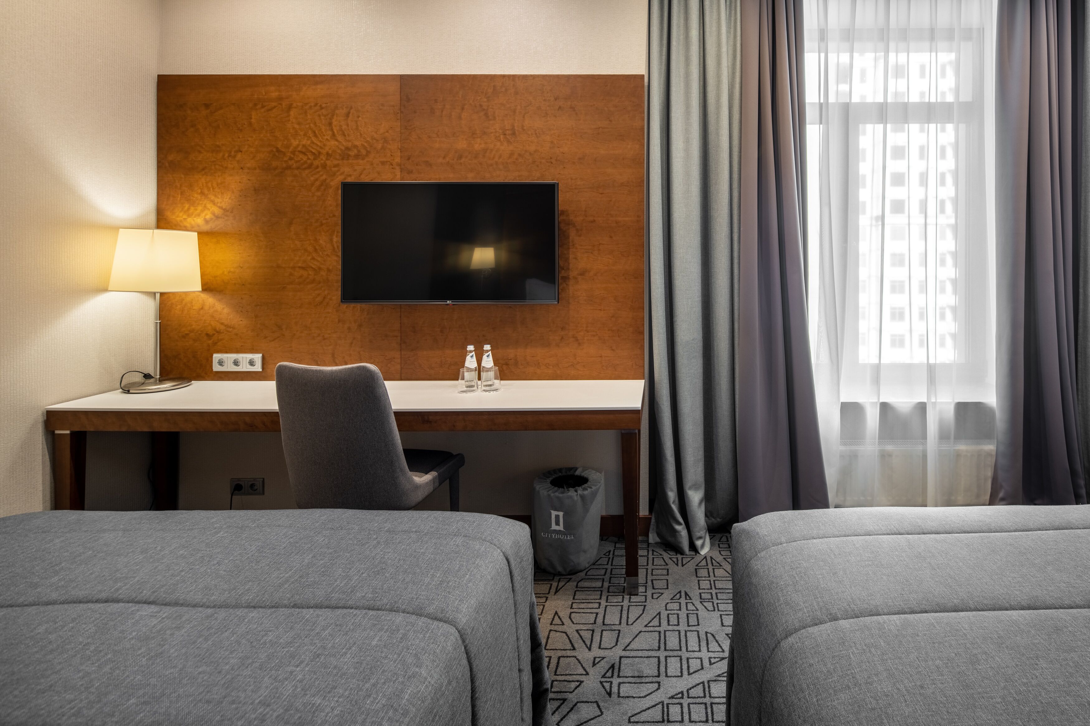 Quarto de hotel com parede de destaque em madeira, televisão de tela plana, escrivaninha com luminária, duas camas e janela com cortinas.