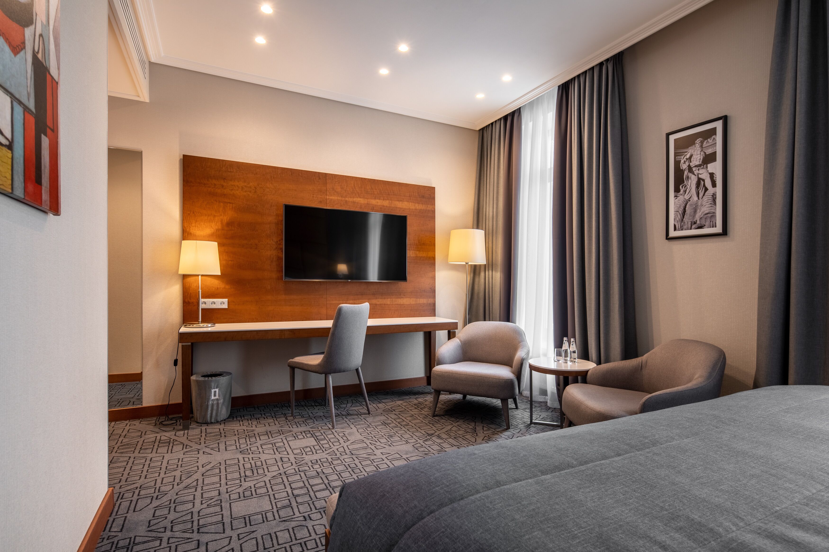 A hotel room with a bed, two armchairs, a desk with a flat-screen TV, a lamp, and a framed picture on the wall.