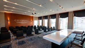 Uma sala de conferências com uma mesa comprida e fileiras de cadeiras, amplas janelas com cortinas e uma parede de madeira com o texto "CITYHOTEL CONFERENCE HALL".