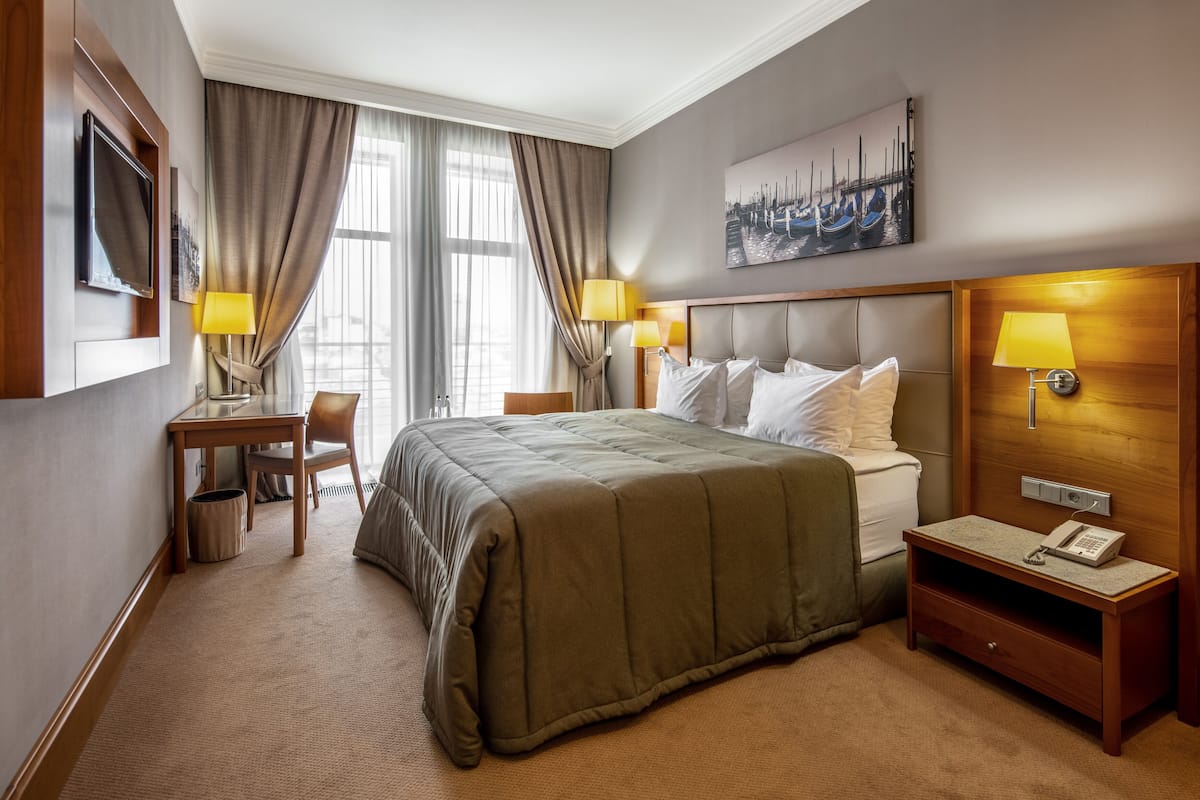 A hotel room with a bed, a desk, a chair, a nightstand with a phone, and a painting on the wall.