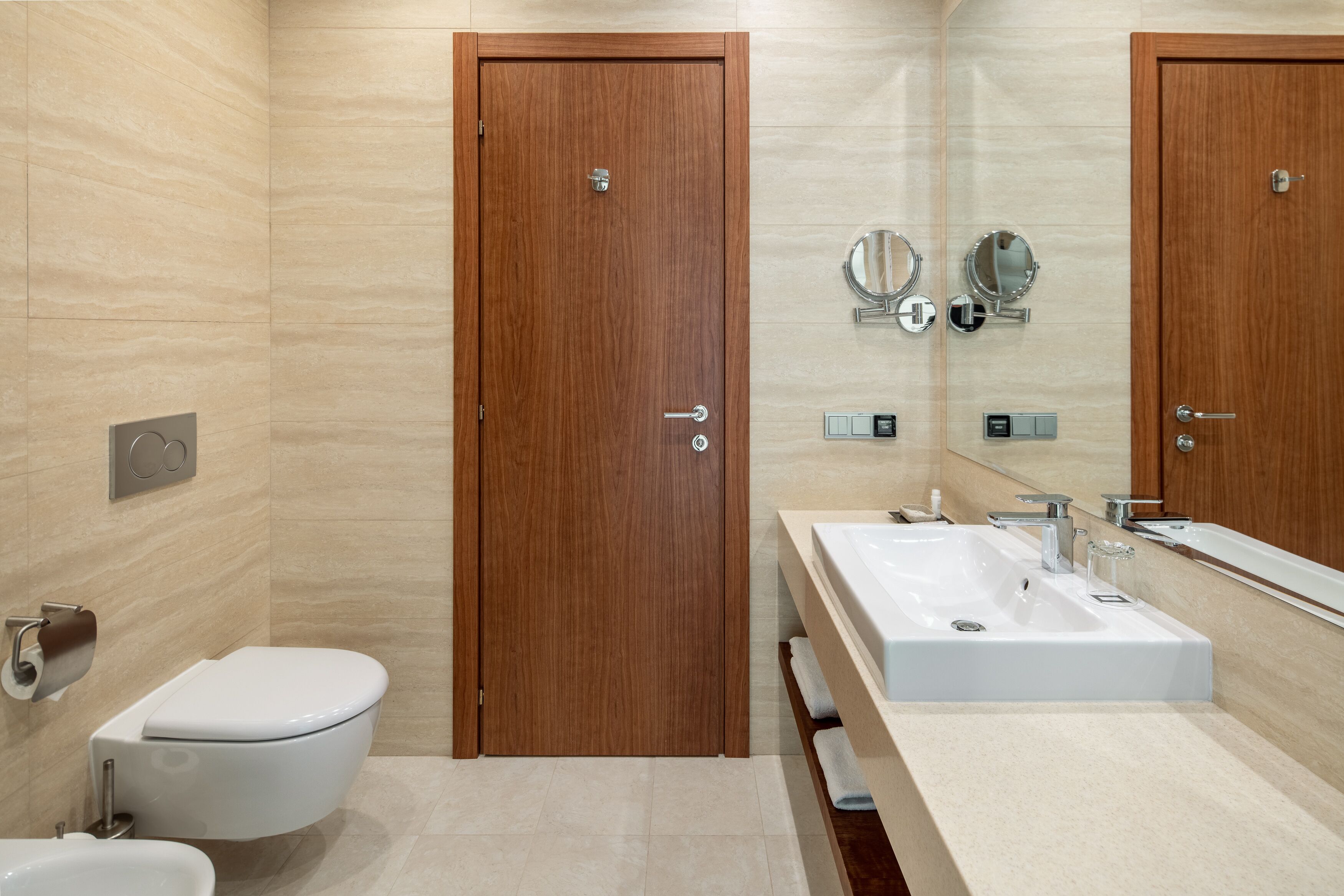 Een moderne badkamer met een houten deur, een wit toilet, een wastafel en twee spiegels aan de muur.