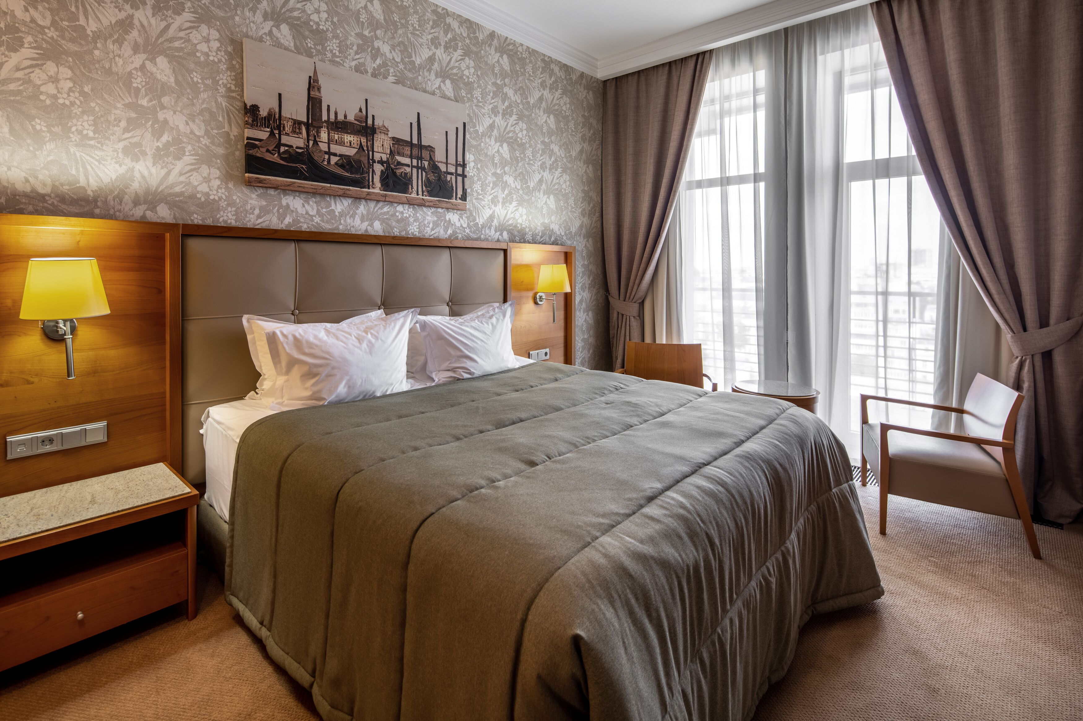 A hotel room with a large bed, two bedside lamps, a wooden nightstand, a chair, and a window with curtains.