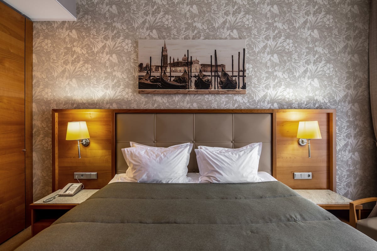 A hotel room with a bed, two bedside lamps, a telephone, and a painting of a cityscape on the wall.