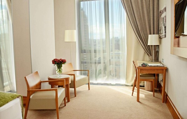 Quarto de hotel com cama, escrivaninha com luminária, duas cadeiras, um vaso com flores e vista para edifícios através de cortinas translúcidas.