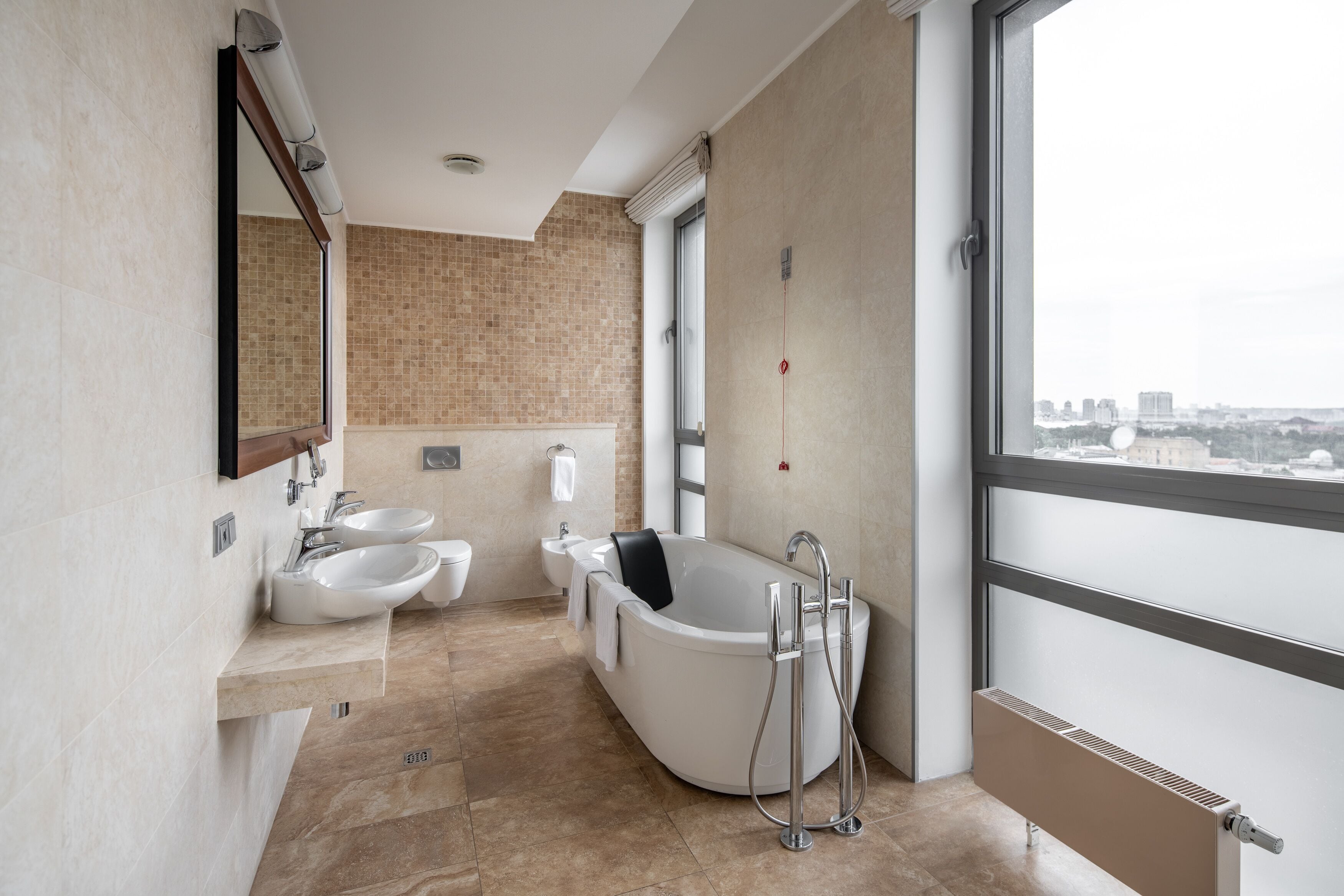 A modern bathroom with a freestanding bathtub, two sinks, and a large window offering a city view.