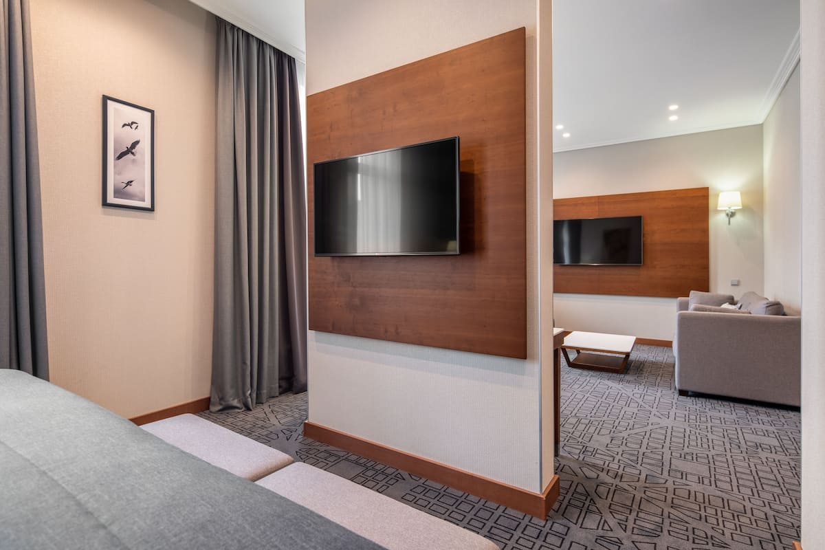 A modern hotel room with a large flat-screen TV mounted on a wooden panel, a grey sofa, a small table, and a patterned carpet.