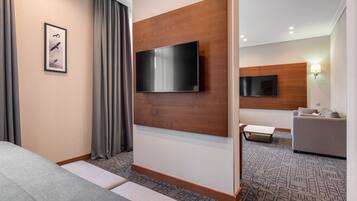 A modern hotel room with a large flat-screen TV mounted on a wooden panel, a grey sofa, a small table, and a patterned carpet.