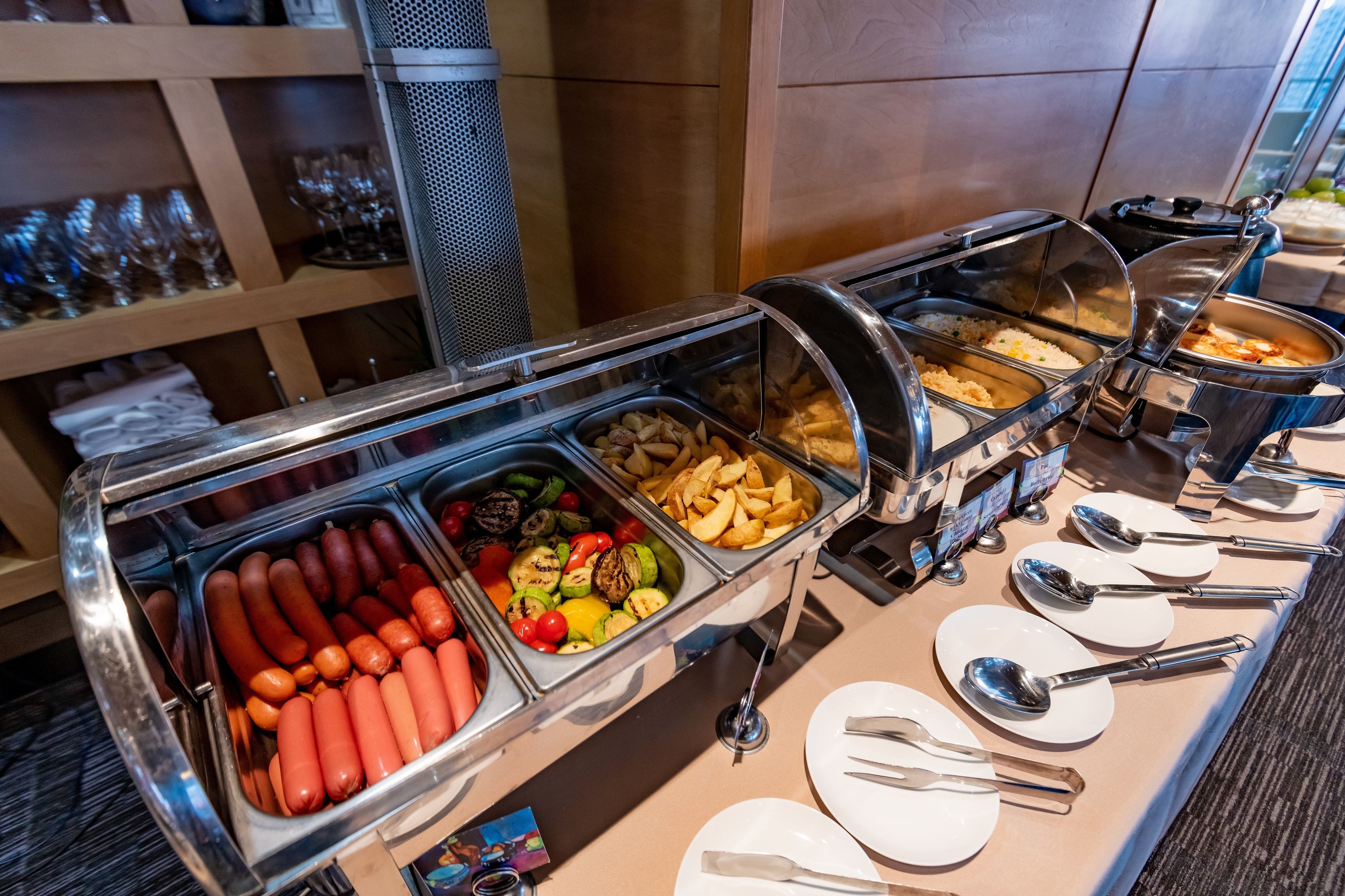Un bufé con diversas opciones de comida, incluyendo salchichas, verduras a la parrilla y papas fritas.