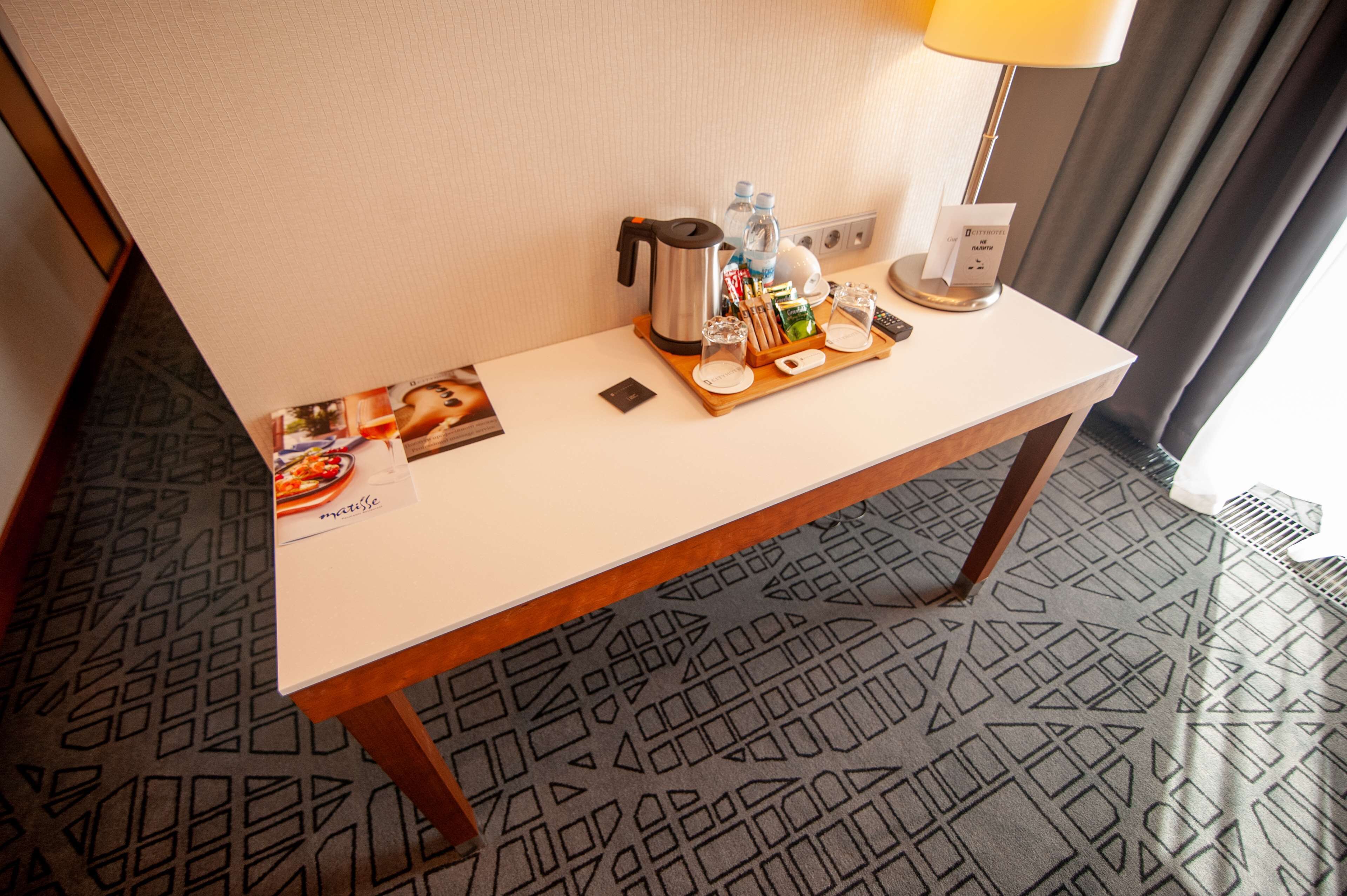 Habitación de hotel con una mesa ordenada que incluye una cafetera, botellas de agua, una bandeja con vasos y aperitivos, y un menú.