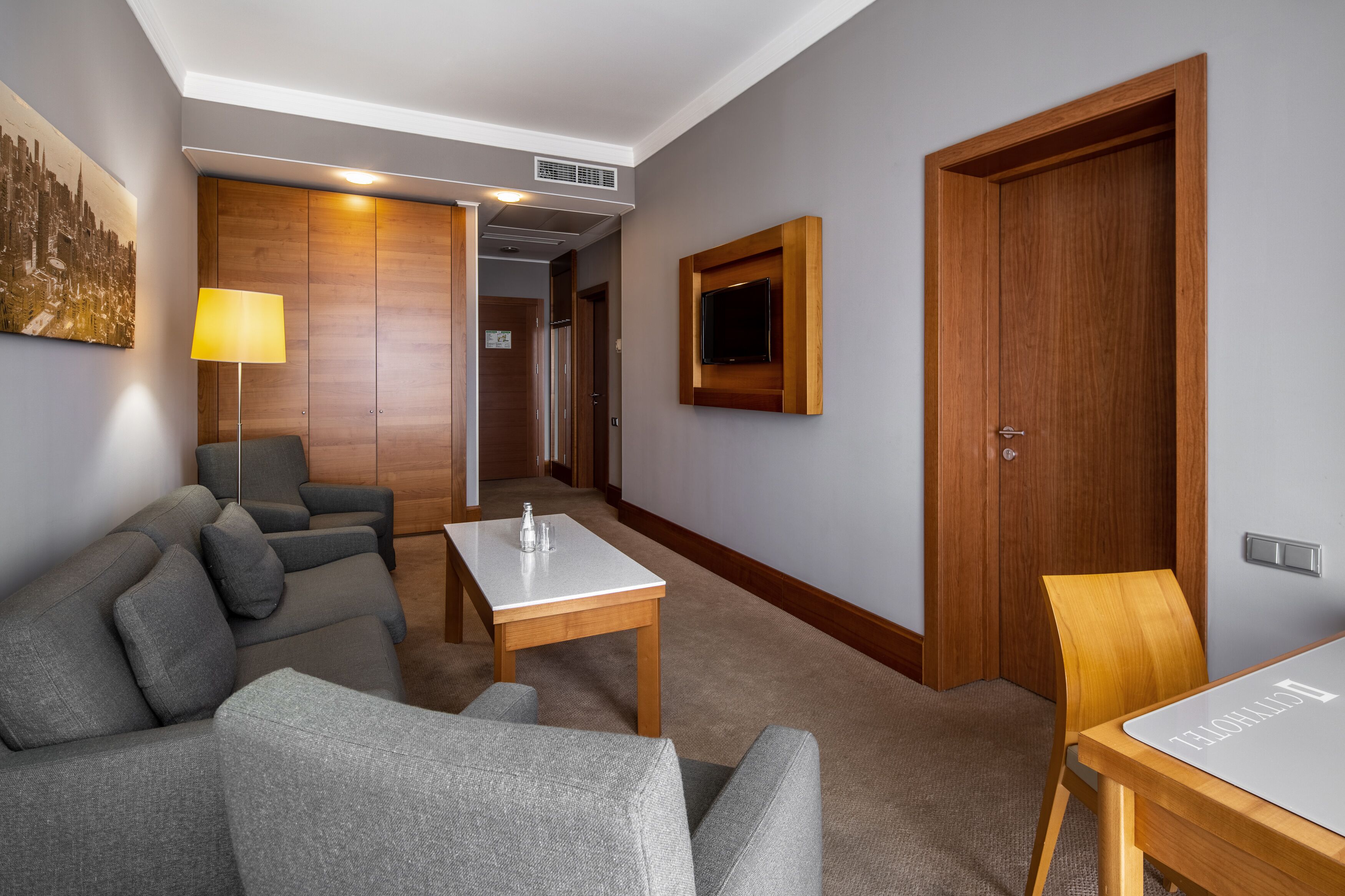 A modern hotel room with a grey sofa, a small table, a wooden door, and a TV mounted on the wall.