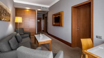 A modern hotel room with a grey sofa, a small table, a wooden door, and a TV mounted on the wall.
