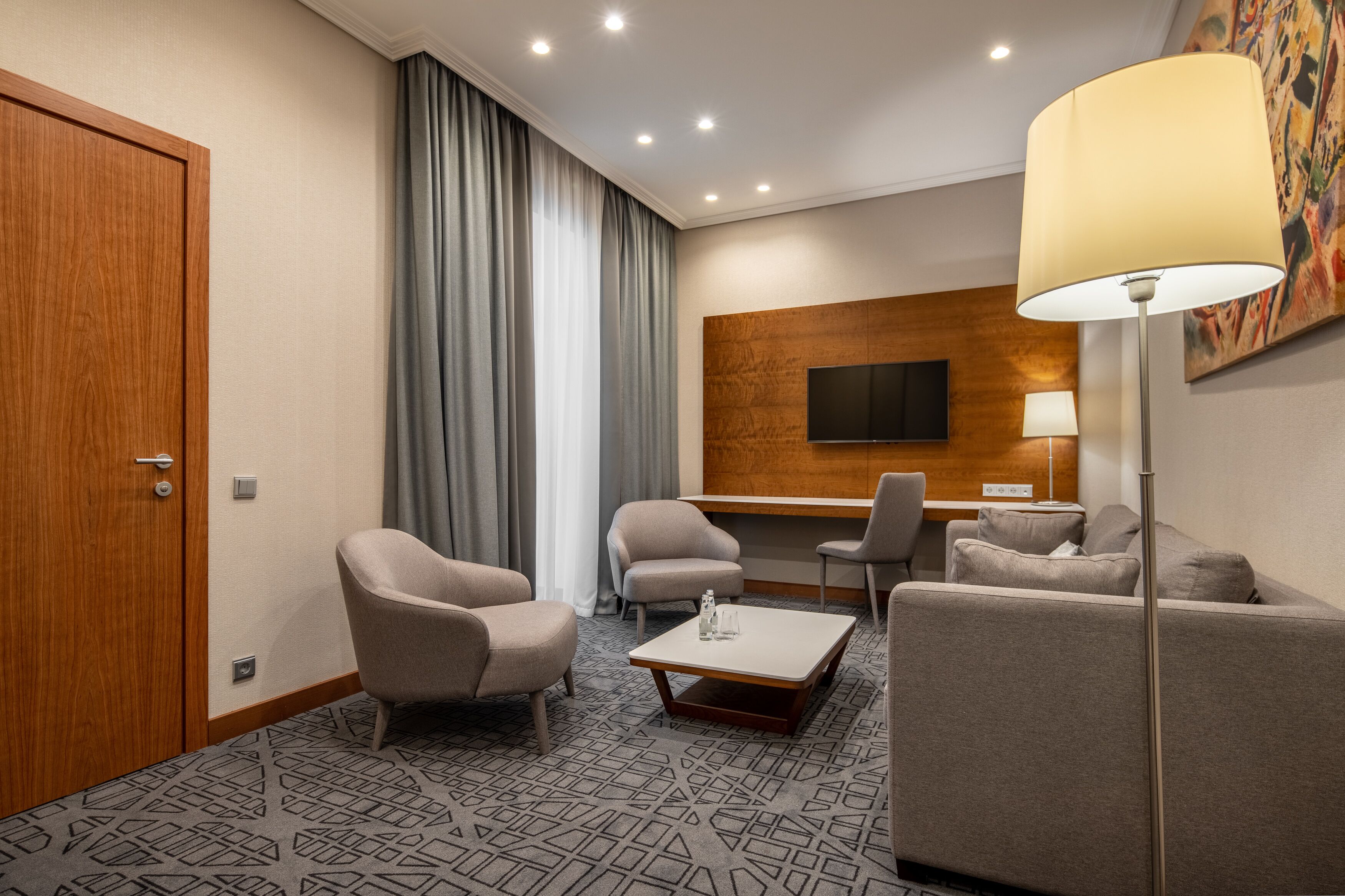 A hotel room with a wooden door, grey curtains, a flat-screen TV mounted on a wooden panel, a desk with a lamp, and two armchairs.