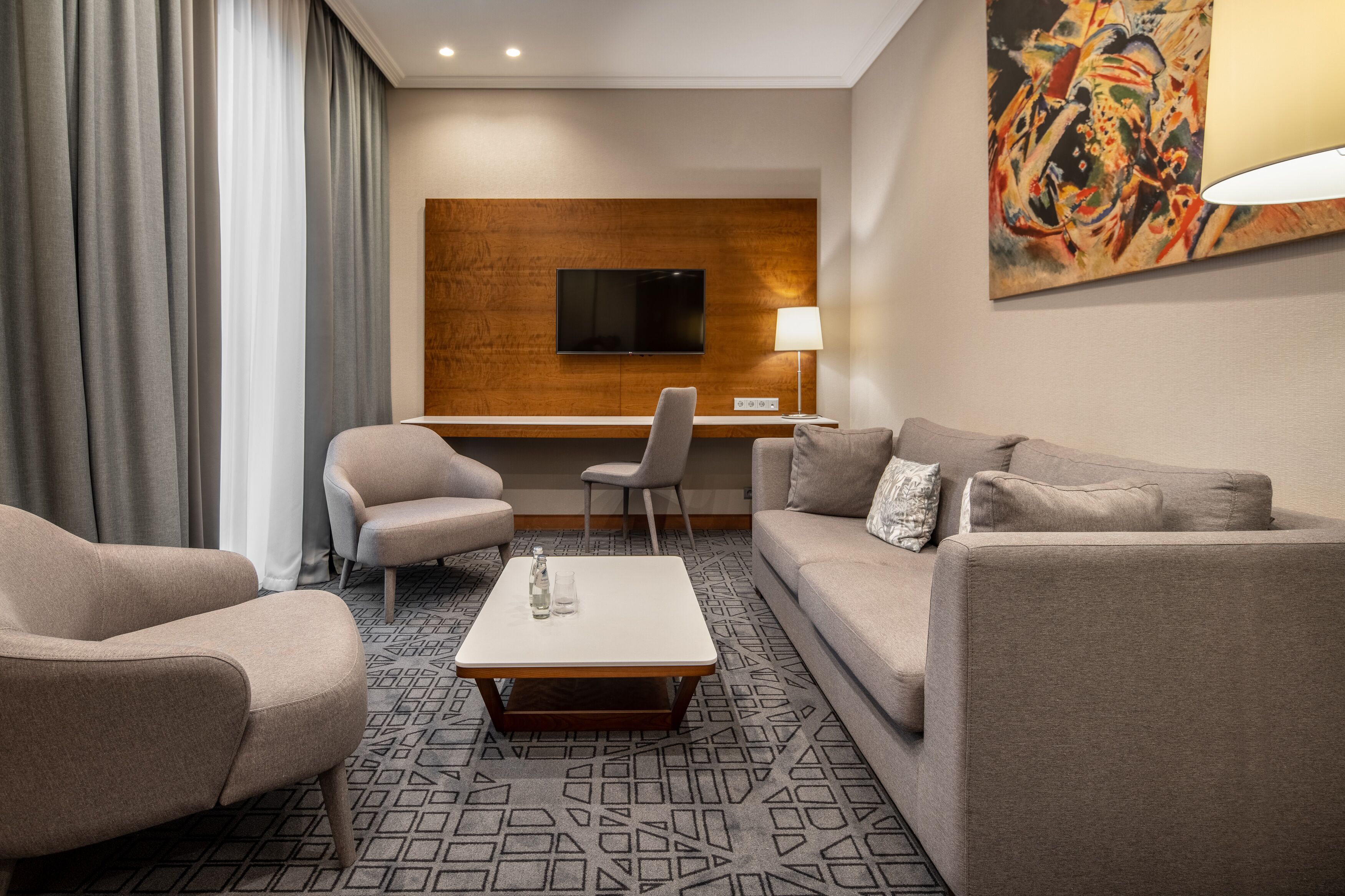 Ein modernes Hotelzimmer mit einer Couch, zwei Sesseln, einem Couchtisch, einem Schreibtisch, einem Flachbildfernseher und einem Bild an der Wand.