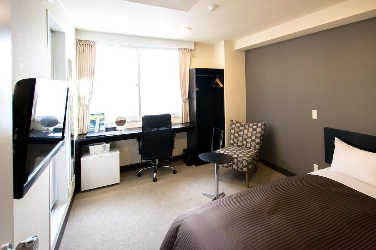 A hotel room with a bed, a desk, a chair, a wardrobe, and a window with curtains.