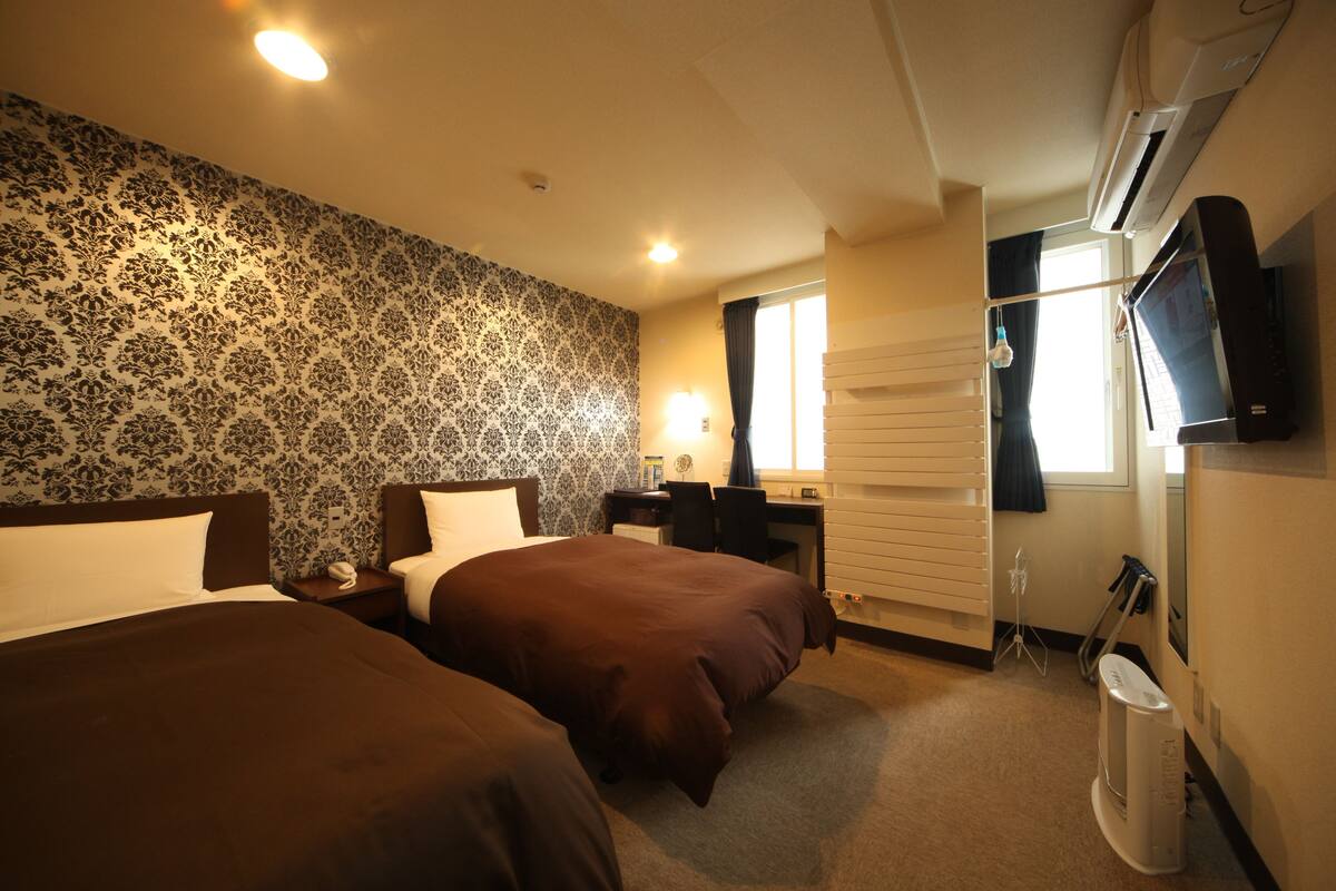A hotel room with two beds, a desk, a chair, a TV, and a window with blinds.