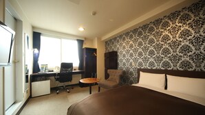 A hotel room with a large bed, a desk with a chair, a small table, and a wardrobe.