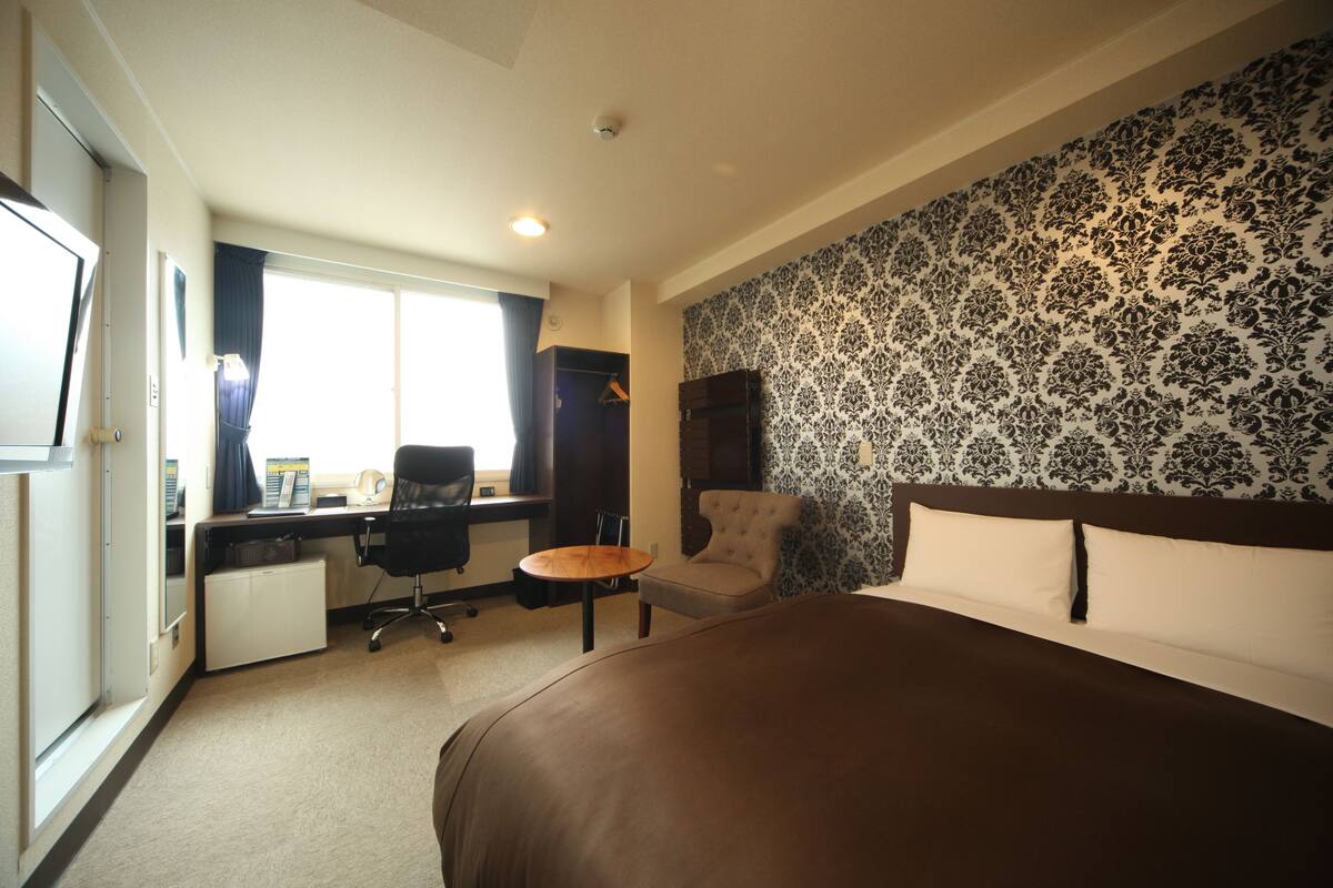 A hotel room with a large bed, a desk with a chair, a small table, and a wardrobe.