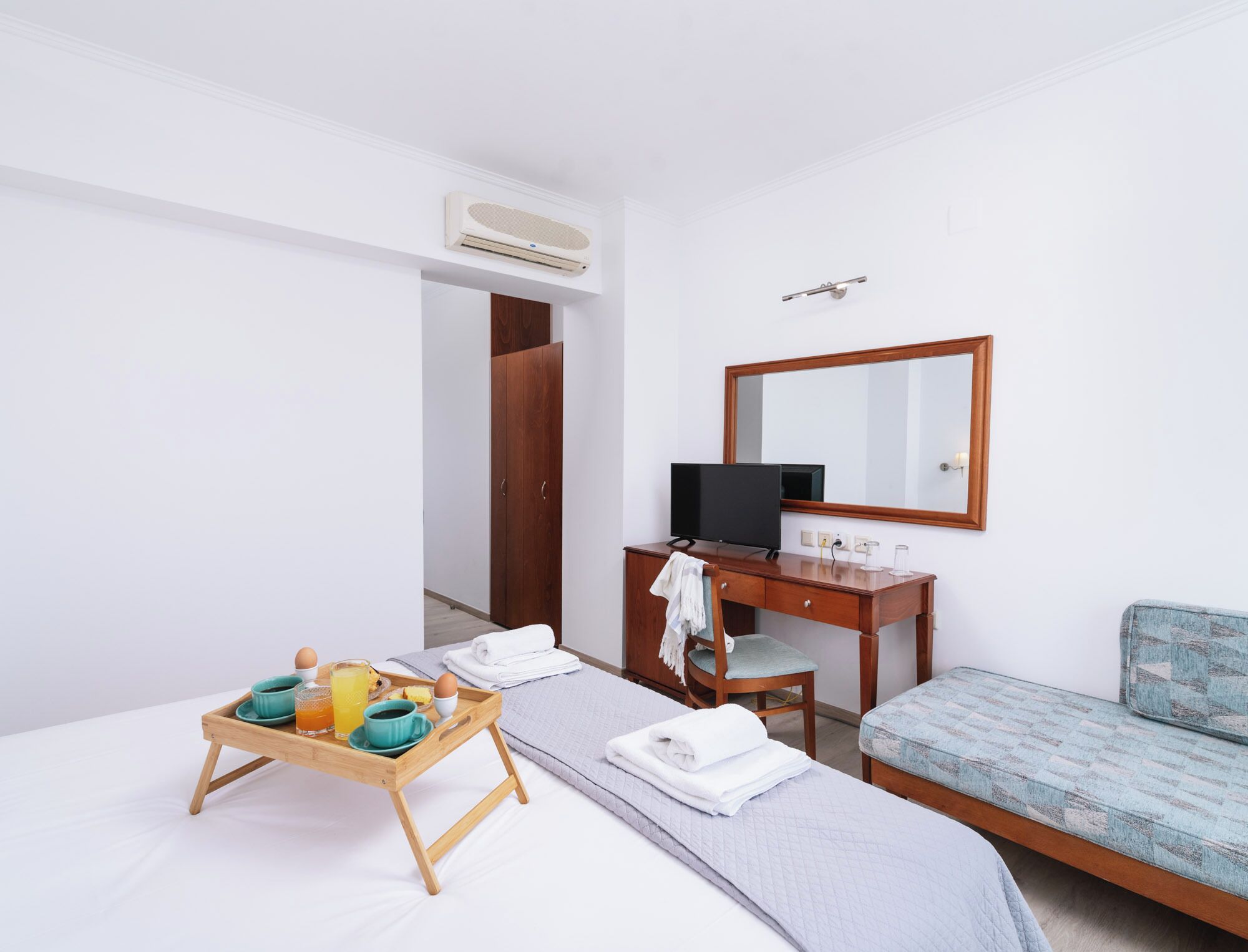 A hotel room with a bed, a desk with a computer, a chair, a small sofa, and a tray with breakfast items.