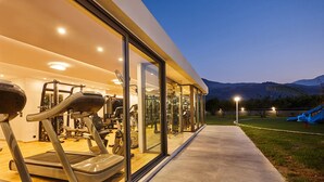 A gym with treadmills and weight machines, a concrete pathway, and a view of a mountain and playground in the background. - Annas House (Apokoronas)
