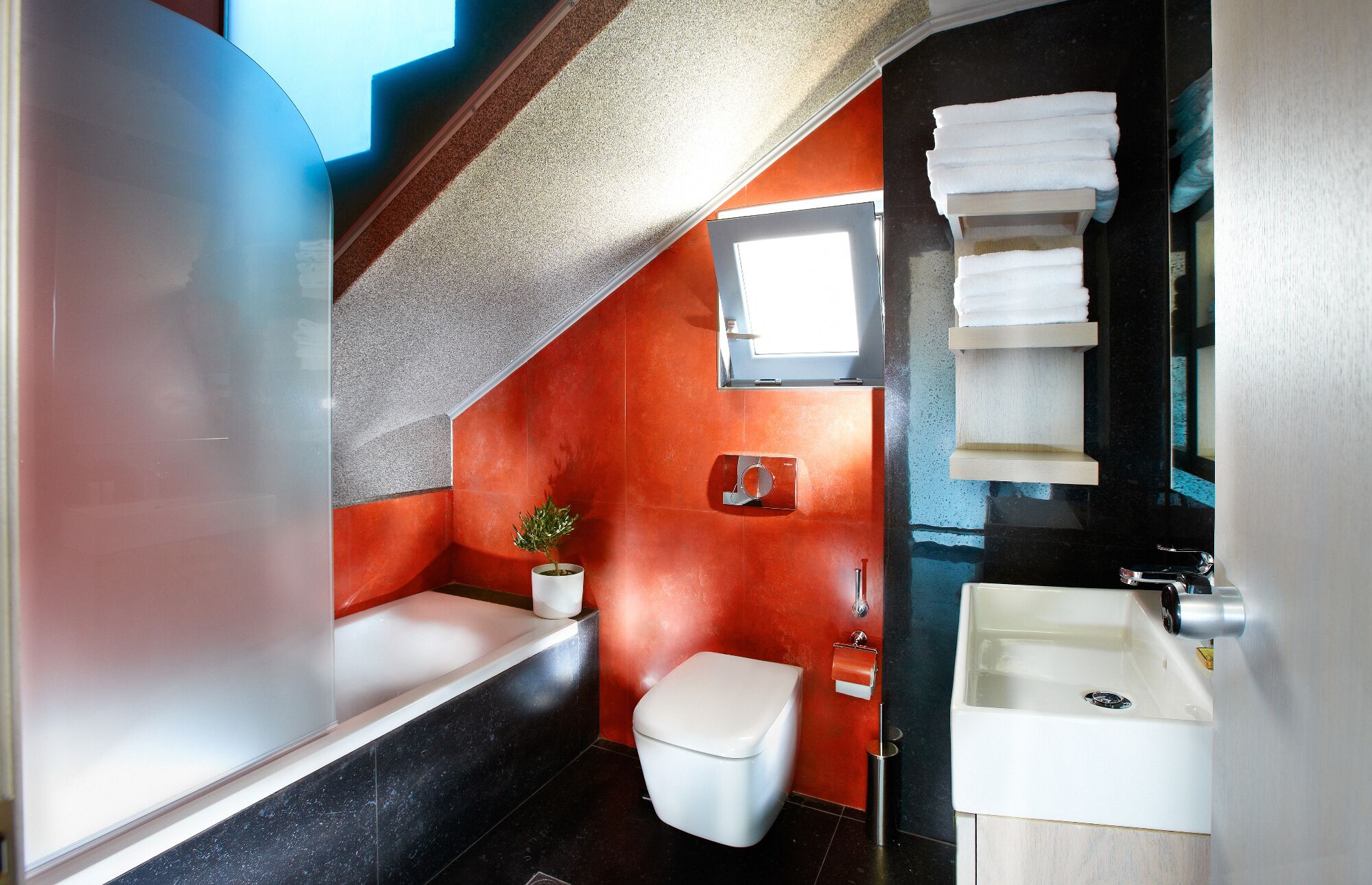 Une salle de bain moderne avec des toilettes blanches, un lavabo et une douche. Une fenêtre laisse entrer la lumière naturelle et une plante est posée sur le sol.