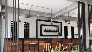 A bar area with a white brick wall, wooden bar counter, stools, hanging pendant lights, and a framed shelf with bottles. - Paragon Inn (Bang Phli)