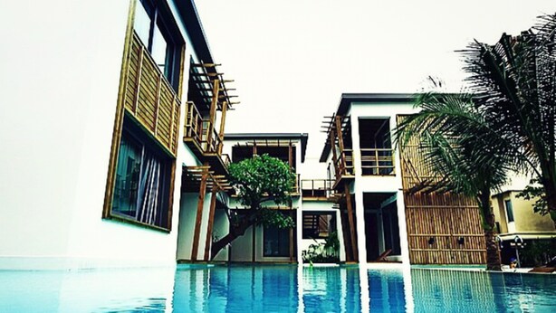 A swimming pool with a reflection of white flowers, surrounded by modern buildings with balconies and a palm tree. - Paragon Inn (Bang Phli)