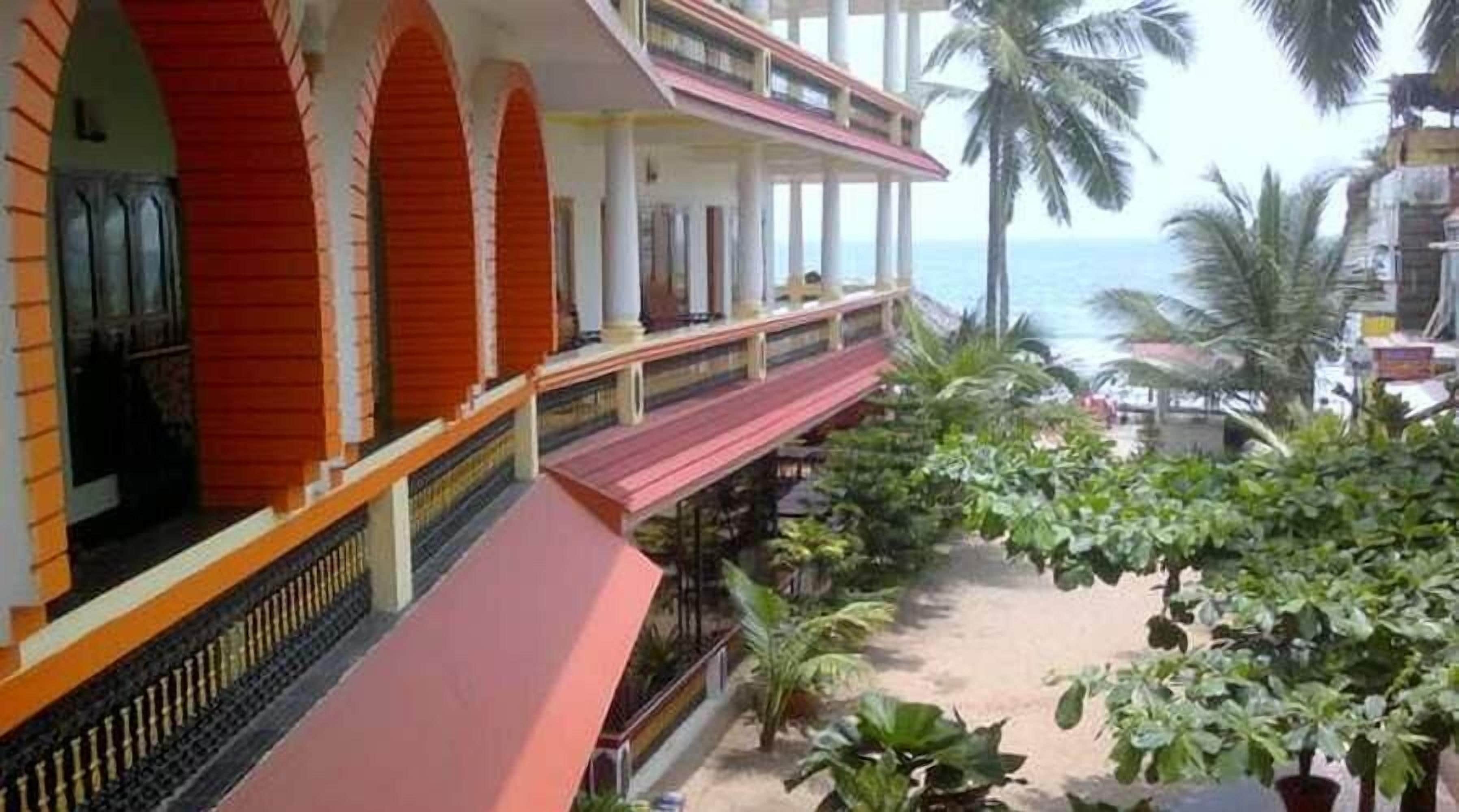 A hotel with a red and white facade, a balcony with a view of the ocean, and tropical plants.