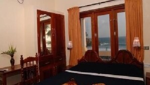 A bedroom with a bed, a wooden desk, a chair, a potted plant, and a view of the ocean through a window.
