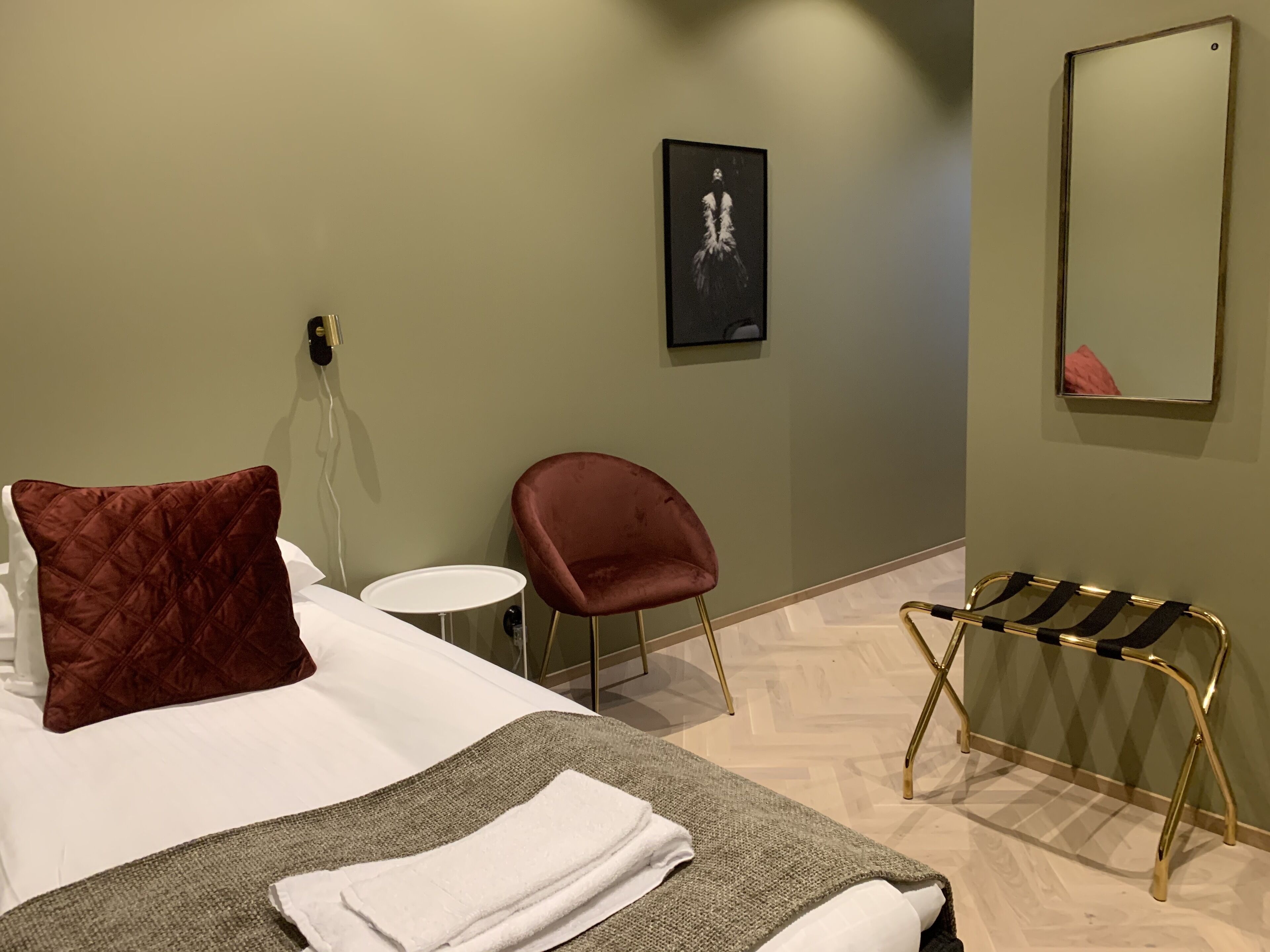 Habitación de hotel con cama, una silla roja, una mesita redonda pequeña, toalla, espejo y un cuadro colgado en la pared.