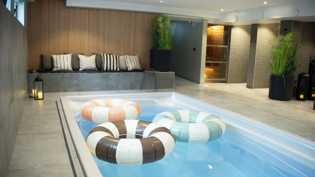 A modern indoor pool with colorful floating rings, a seating area with cushions, and potted plants. - Hotell Esplanad (Växjö)