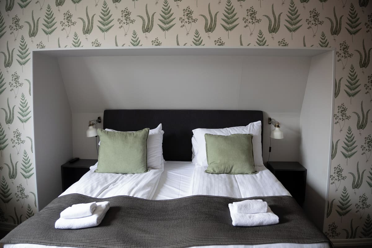 A double bed with white linens and two green pillows, flanked by nightstands with lamps, in a room with fern-patterned wallpaper.