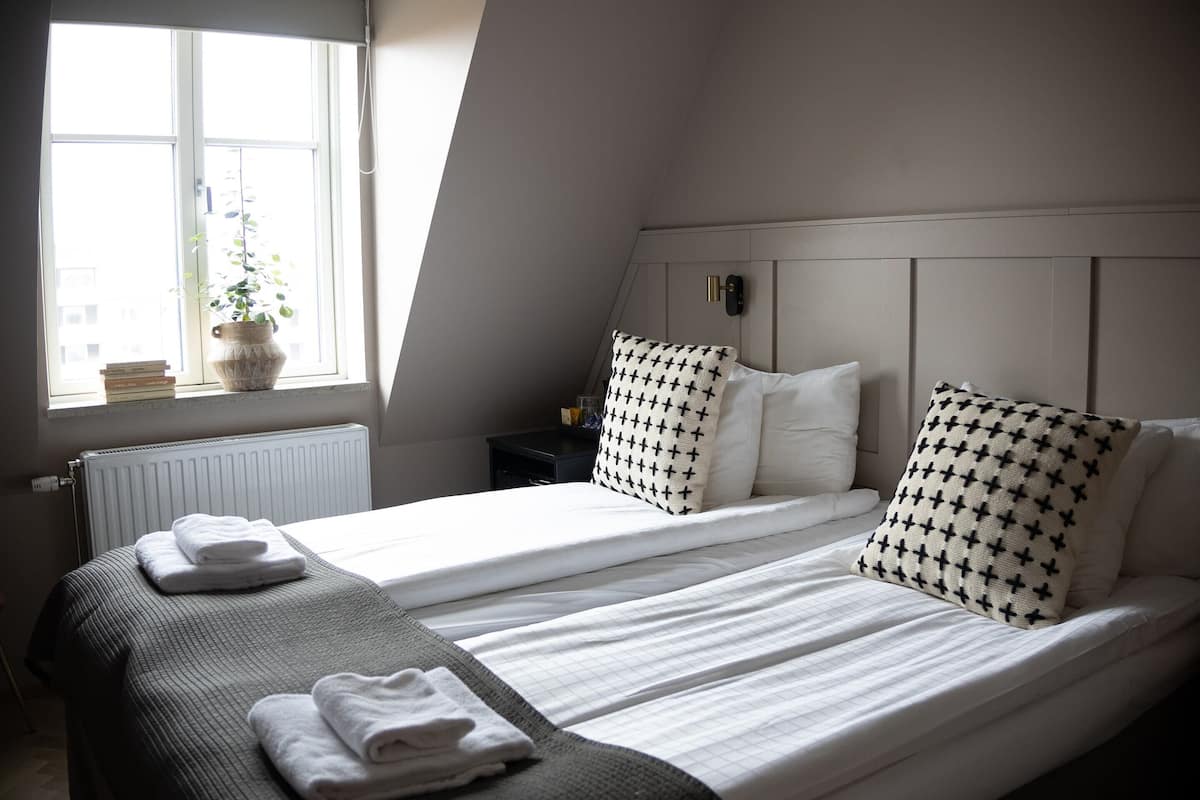 A hotel room with two beds, a window with a view, a radiator, and a bedside table with a lamp.