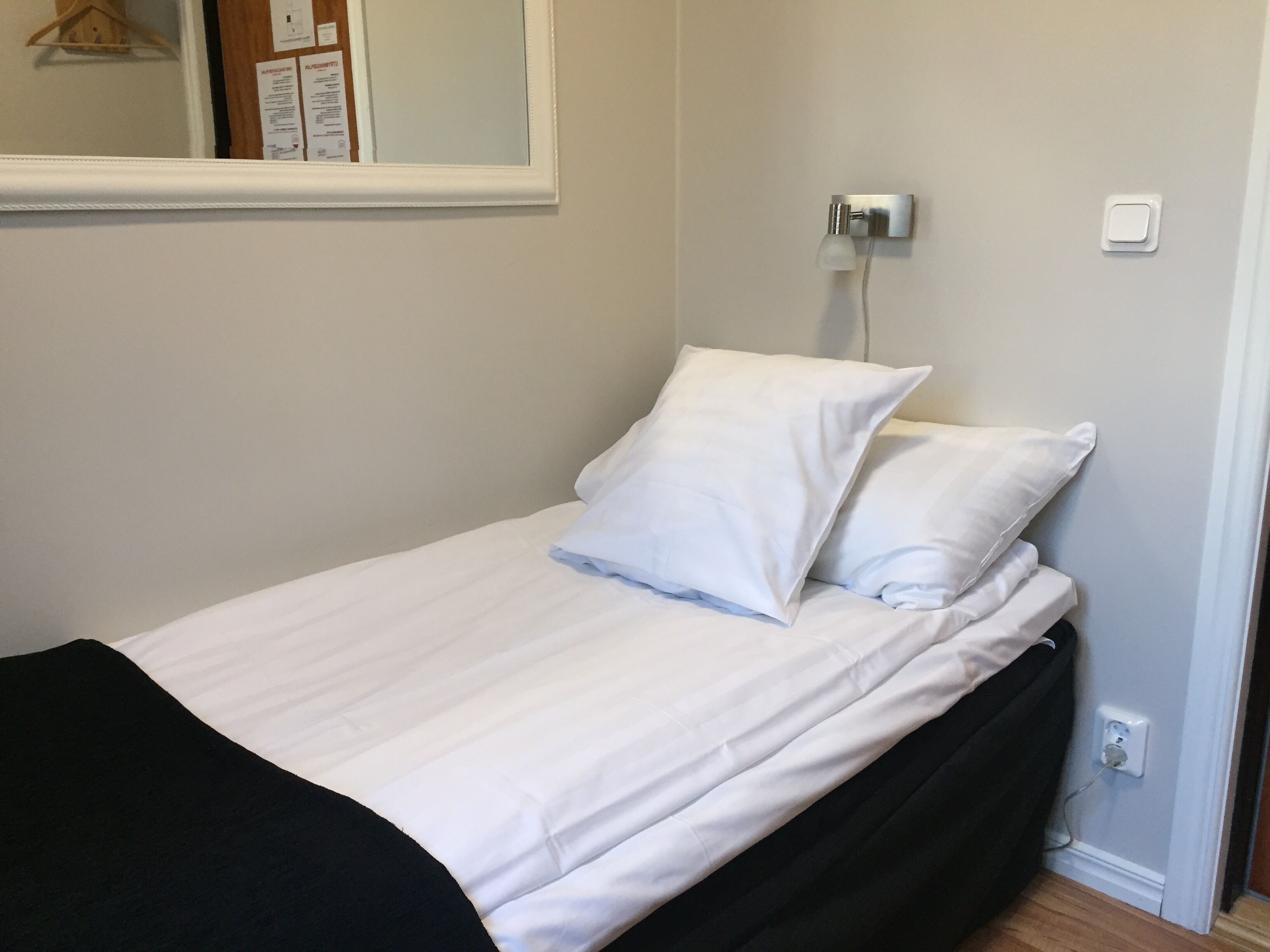 Una cama pequeña y bien hecha con sábanas blancas y dos almohadas, una mesita de noche con lámpara y un enchufe en la pared.