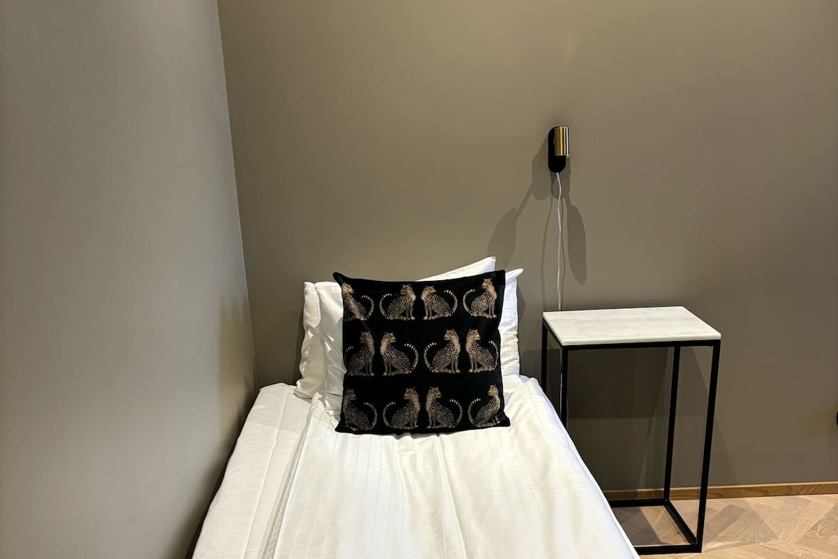 A single bed with white bedding, a patterned black pillow, and a brown throw blanket. A small white nightstand with a black frame is beside the bed.