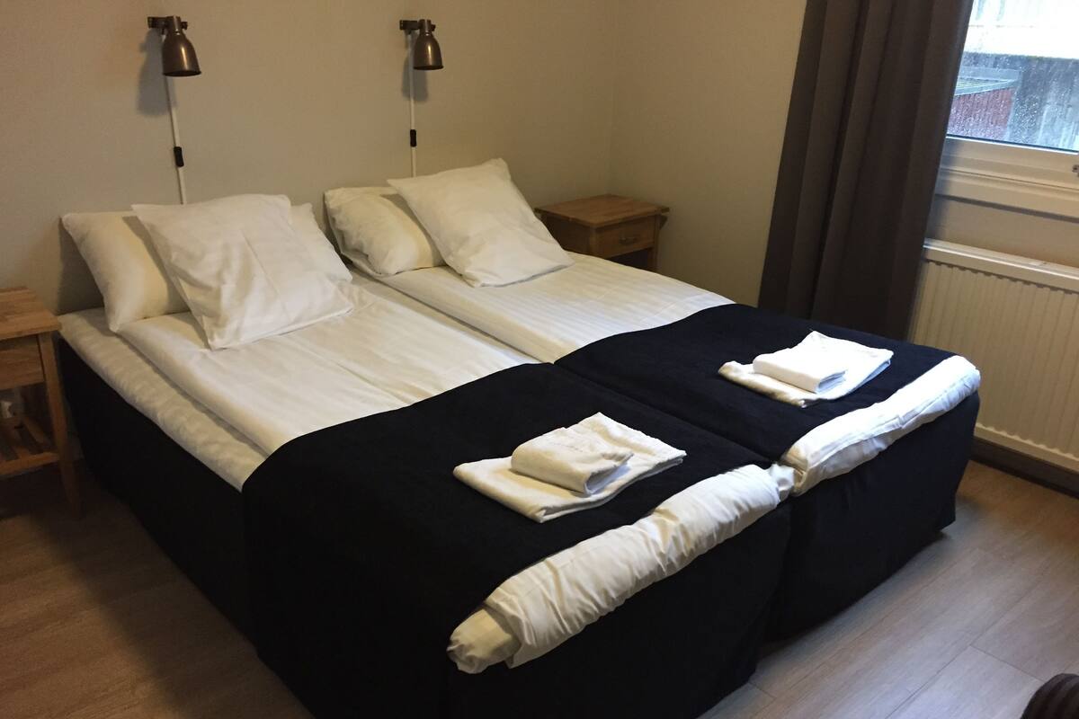A double bed with white linens, a black bedspread, two bedside tables, and a window with curtains.
