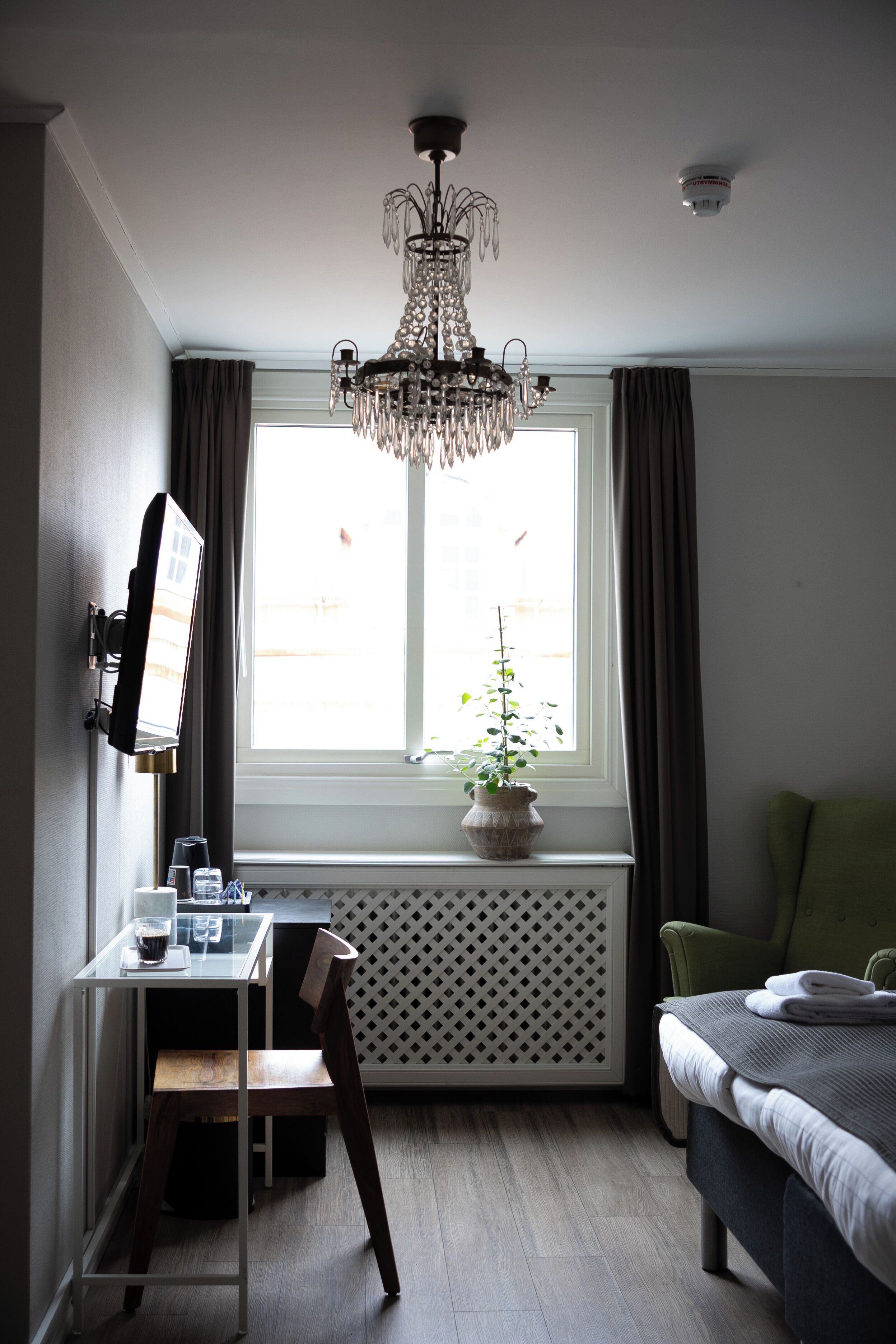 A hotel room with a desk, chair, and a bed. There is a window with curtains, a radiator, and a potted plant.