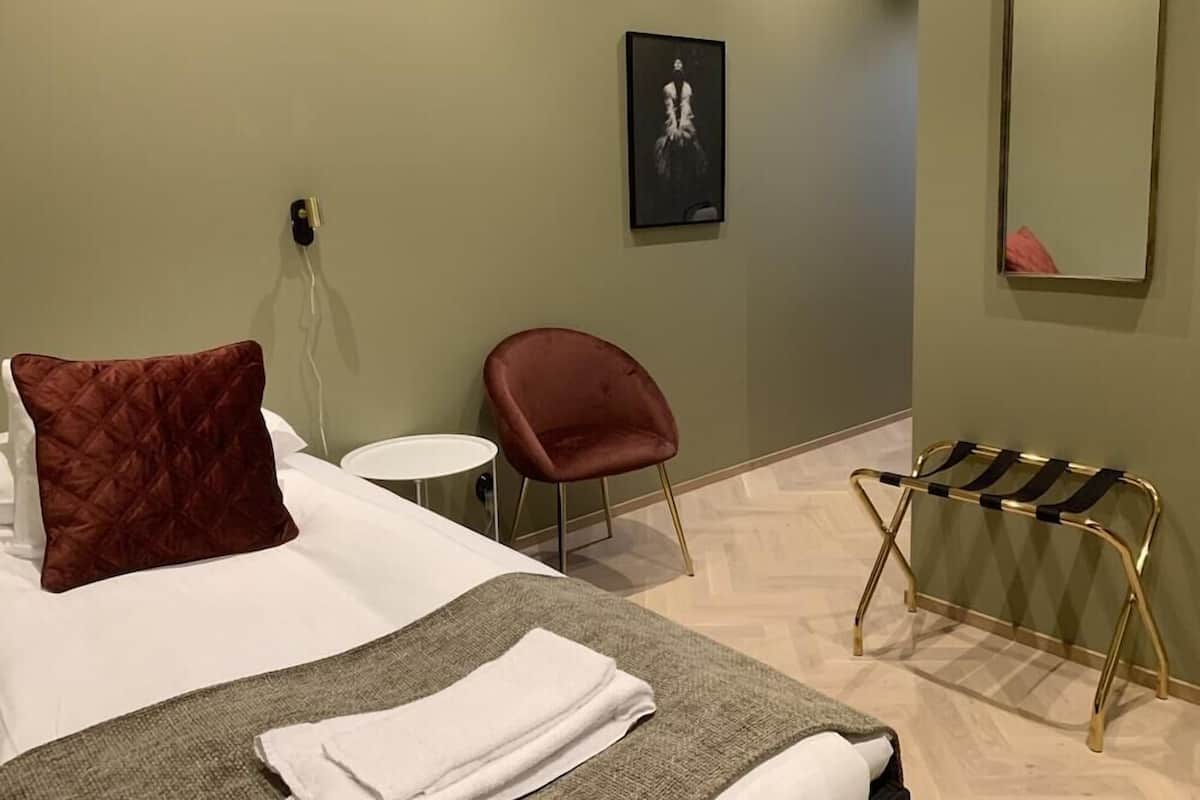 A hotel room with a bed, a red chair, a small round table, a towel, a mirror, and a wall-mounted picture.