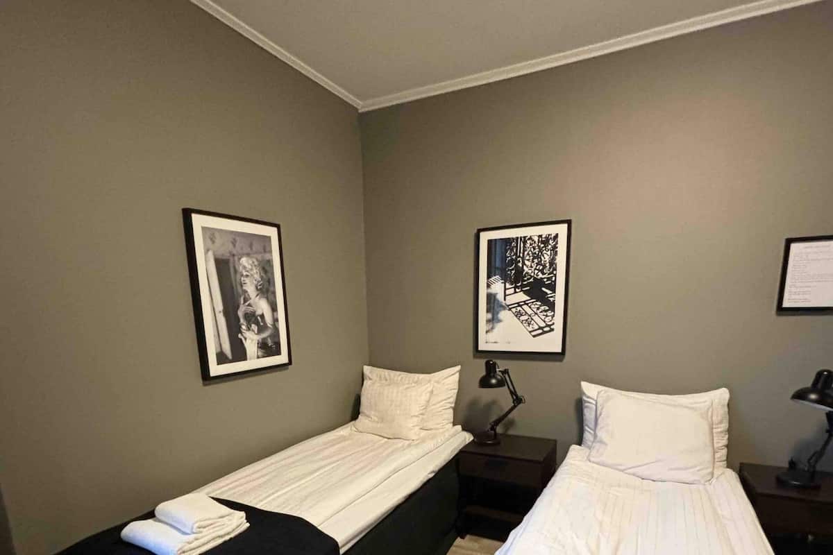 A room with two single beds, each with a black bedspread and white pillows, and a small bedside table with a lamp. There are framed pictures on the walls.