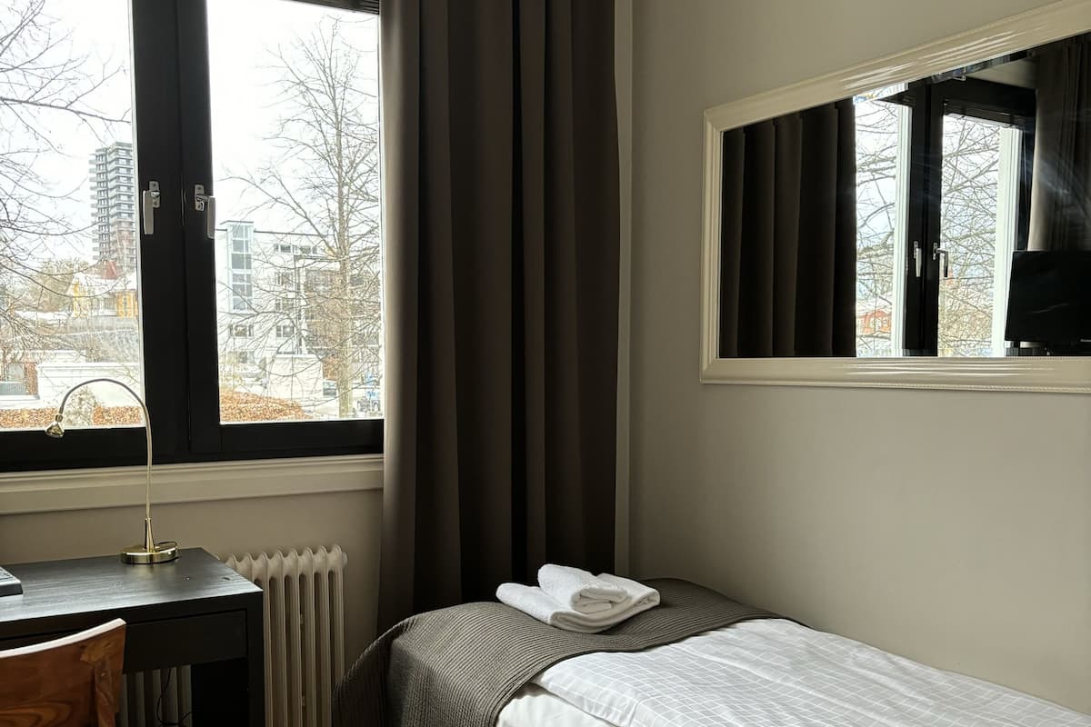 A small, tidy room with a single bed, a desk, and a chair. There is a window with curtains, a radiator, and a view of buildings outside.