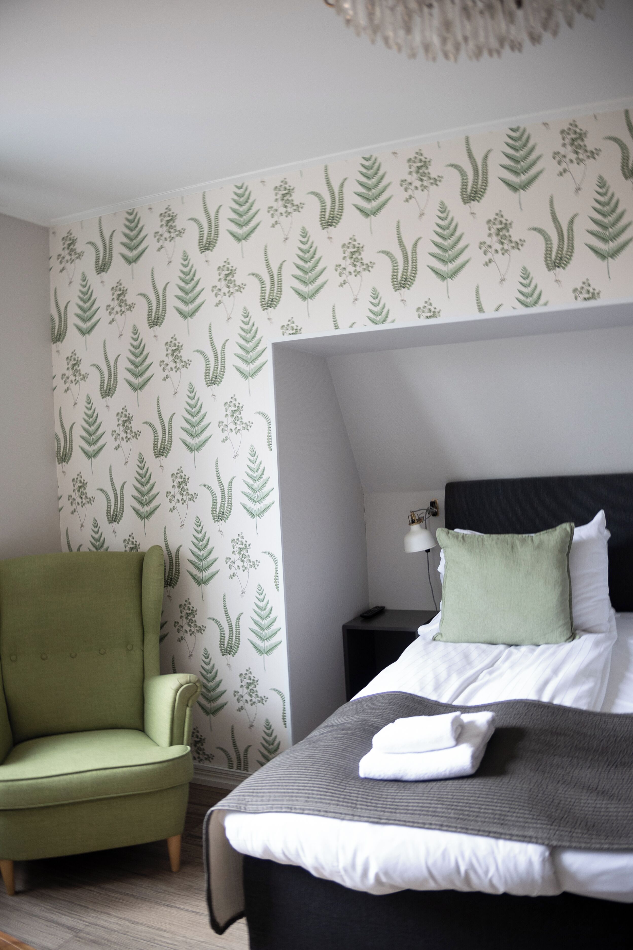 A neatly arranged bedroom with a bed, a green armchair, a nightstand with a lamp, and a patterned wallpaper.