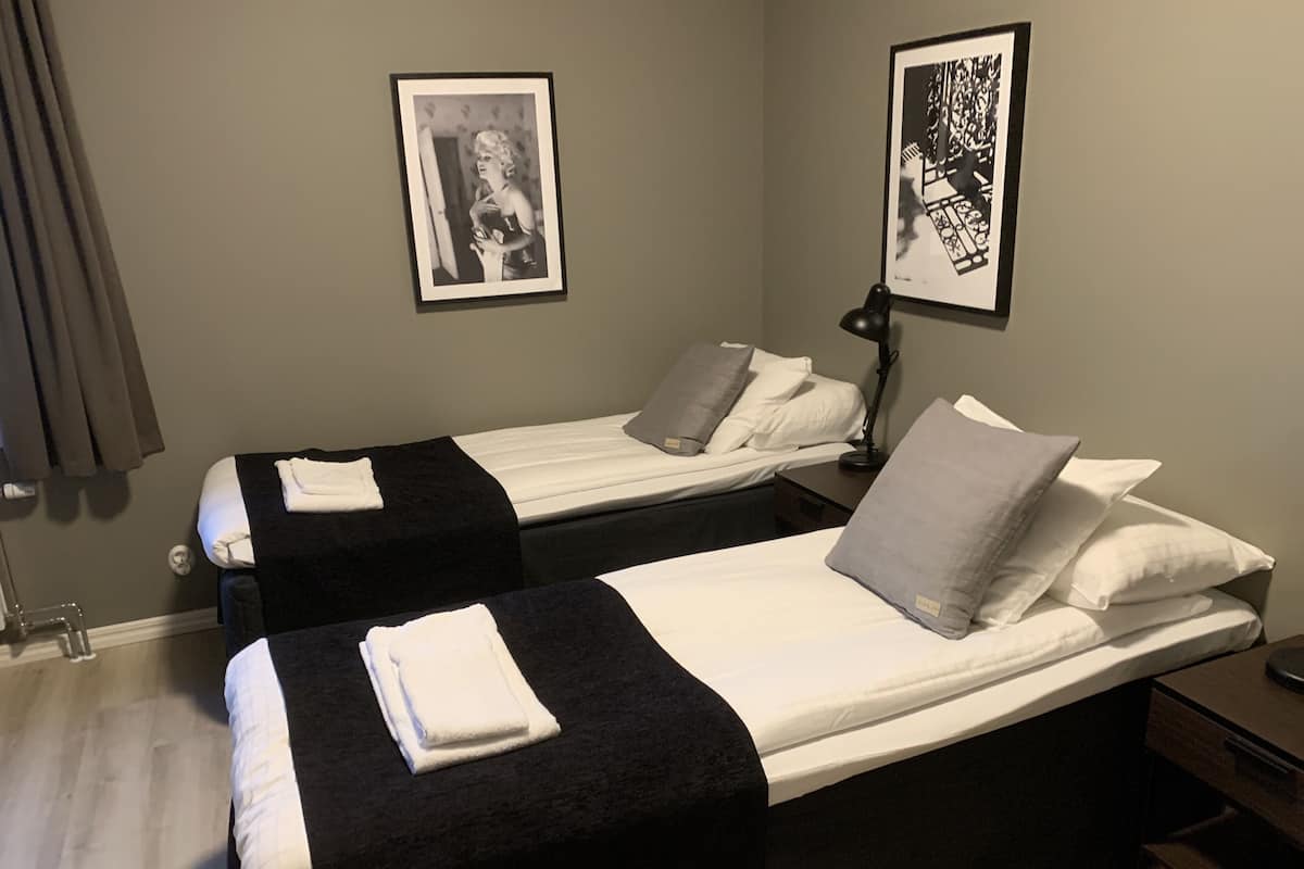 A hotel room with two beds, each with a black bedspread and white pillows, a bedside table with a lamp, and two framed pictures on the wall.