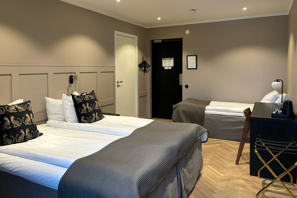 A hotel room with two beds, a desk with a chair, a coat rack, and a door with a sign.