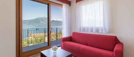 A room with a red sofa, a small table with a vase, a sliding glass door to a balcony with a view of mountains and water.