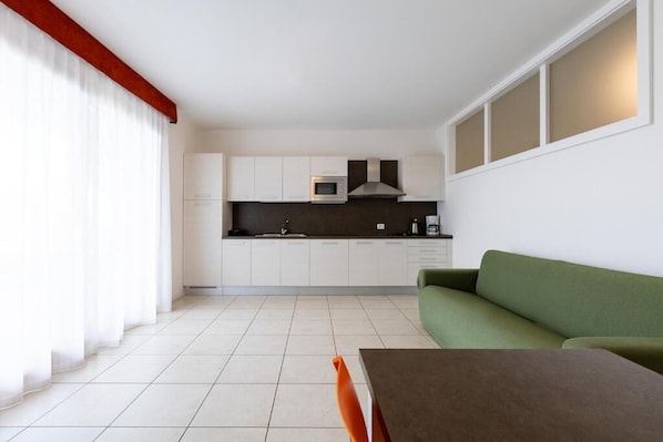 A modern kitchen with white cabinets, a microwave, and a stove. There is a green sofa and a dining table with orange chairs in the living area. - Residence La Corte Danese (Torri del Benaco)