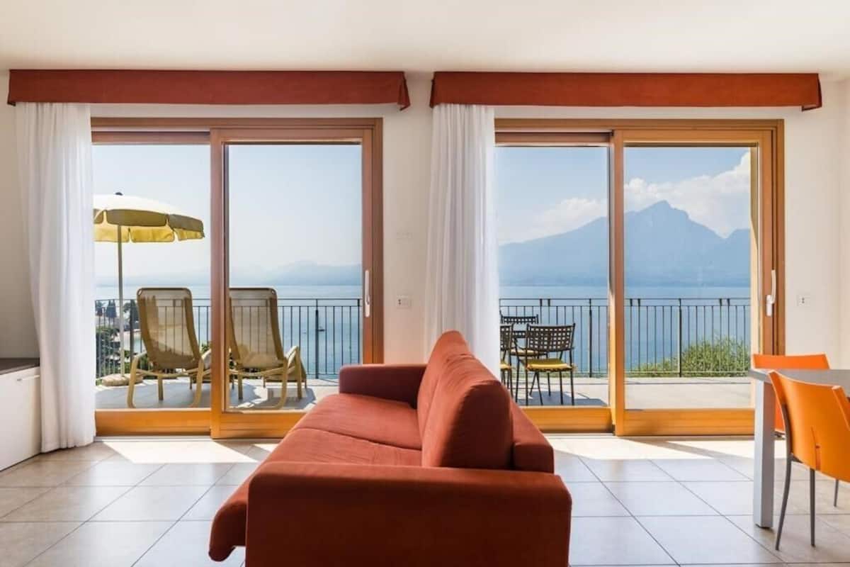 A room with a red sofa, a dining table with orange chairs, and a balcony with chairs and an umbrella.